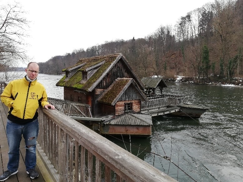 Roman Flassig in gelber Jacke steht im Winter an einem Geländer aus Holz. Dahinter ist ein altes Gebäude aus Holz, das direkt auf dem Wasser gebaut wurde