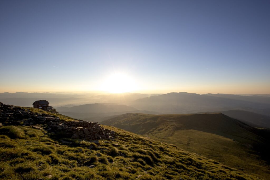 die steirische Berglandschaft ist zu sehen: grüne, grasbewachsene Hügel, dahinter geht die Sonne unter. Man sieht den Zirbitzkogel