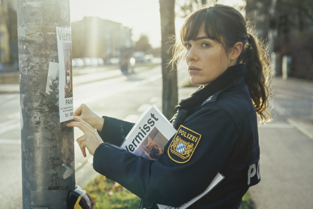Eine Polizistin in Uniform hängt ein Poster auf einem Laternenmasten auf. Darauf steht: Vermisst! Und das Bild einer Katze ist zu sehen
