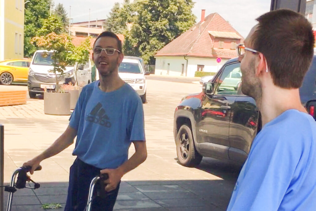 Kevin Neubauer betrachtet sein Spiegelbild in einer großen Scheibe. Er trägt Brille, ein blaues Adidas-T-Shirt und benutzt eine Geh-Hilfe