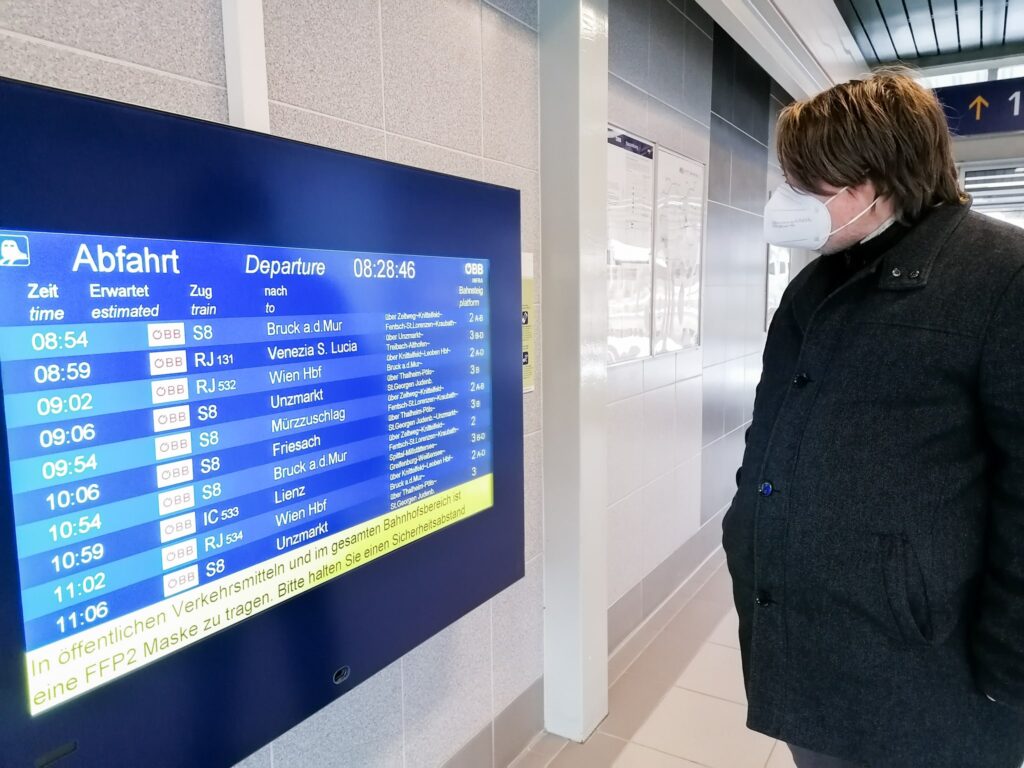 Daniel Gamweger steht am Bahnhof vor einer Anzeigetafel und liest, welche Züge gerade abfahren. Er trägt eine Winterjacke und eine FFP2-Maske