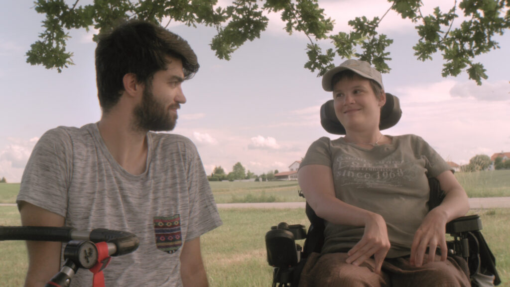 Eva-Maria Proßegger und ihr Assistent Lukas Ladner sitzen im Freien unter einem Baum. Eva-Maria sitzt in ihrem Rollstuhl