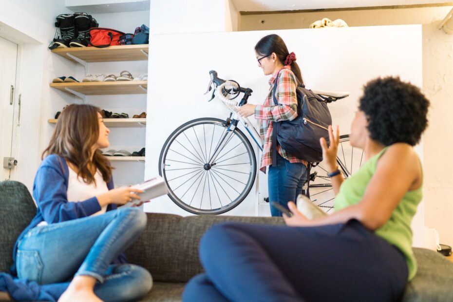 Drei Personen in einem Wohnzimmer, eine steht mit einem Fahrrad, zwei sitzen auf einem Sofa und unterhalten sich.